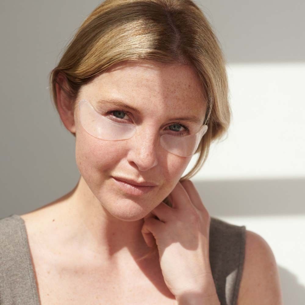 Donna con cuscinetti sotto gli occhi. Indossa un top grigio. Luce solare sullo sfondo.