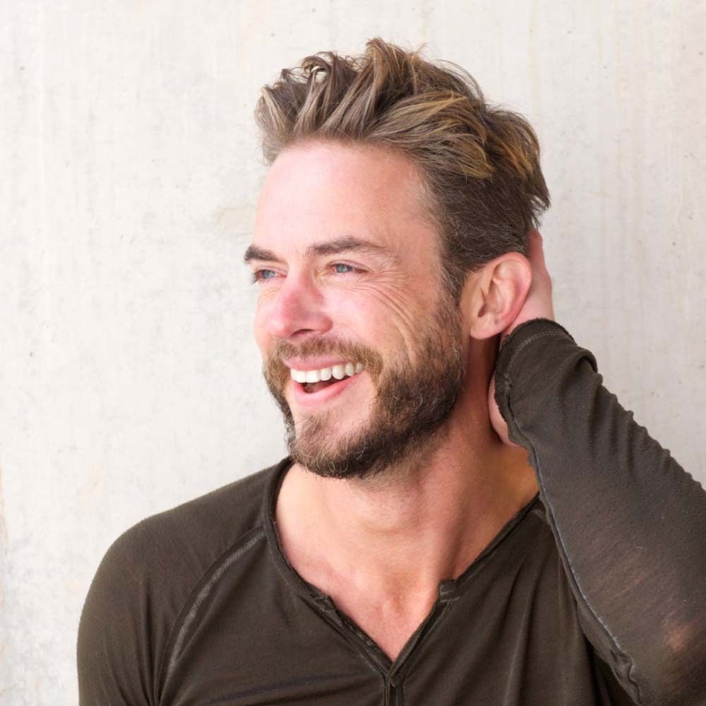 Un uomo con la barba sorride. Si tocca la nuca.