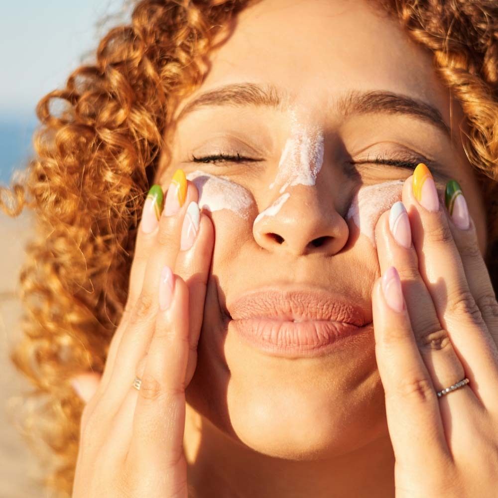 Donna applica crema solare sul viso. Pelle chiara, capelli ricci, unghie colorate. Primo piano.