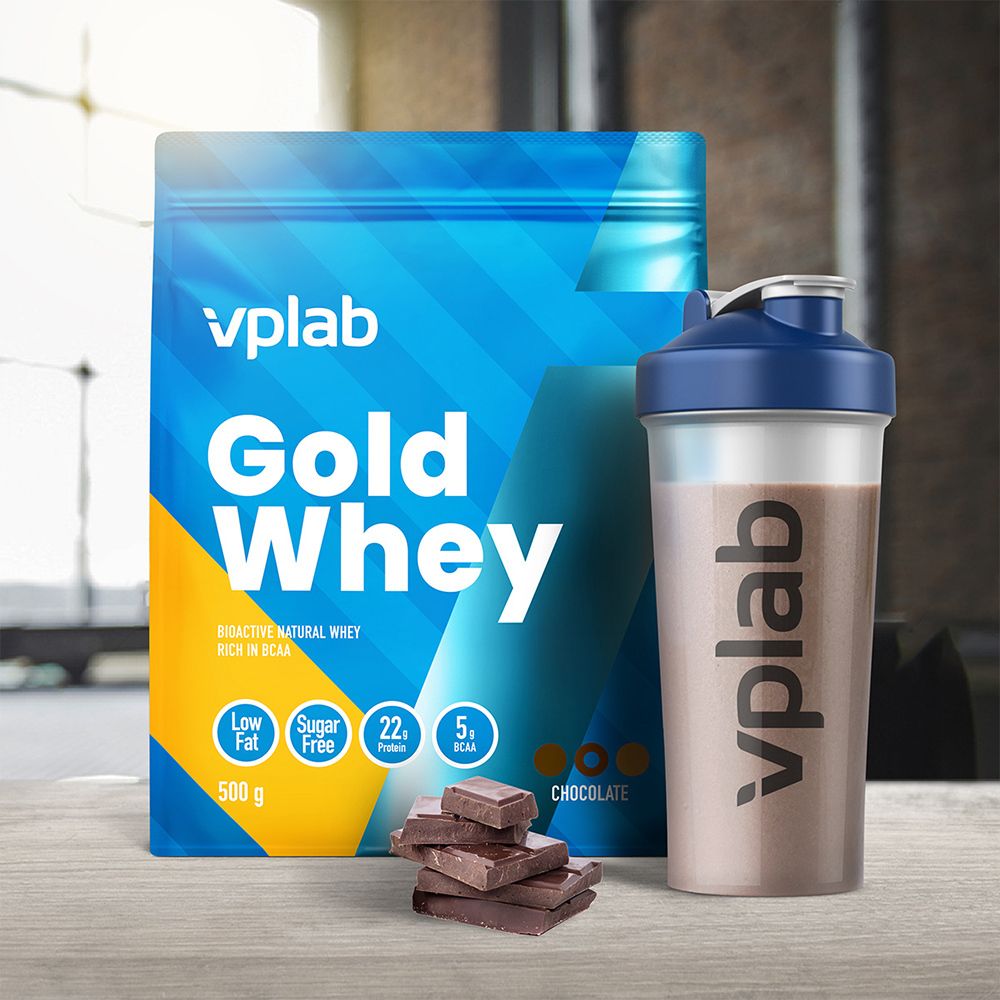 Confezione blu 'Gold Whey', shaker e pezzi di cioccolato. Shaker con logo 'vplab'. Prodotto accanto al cioccolato.
