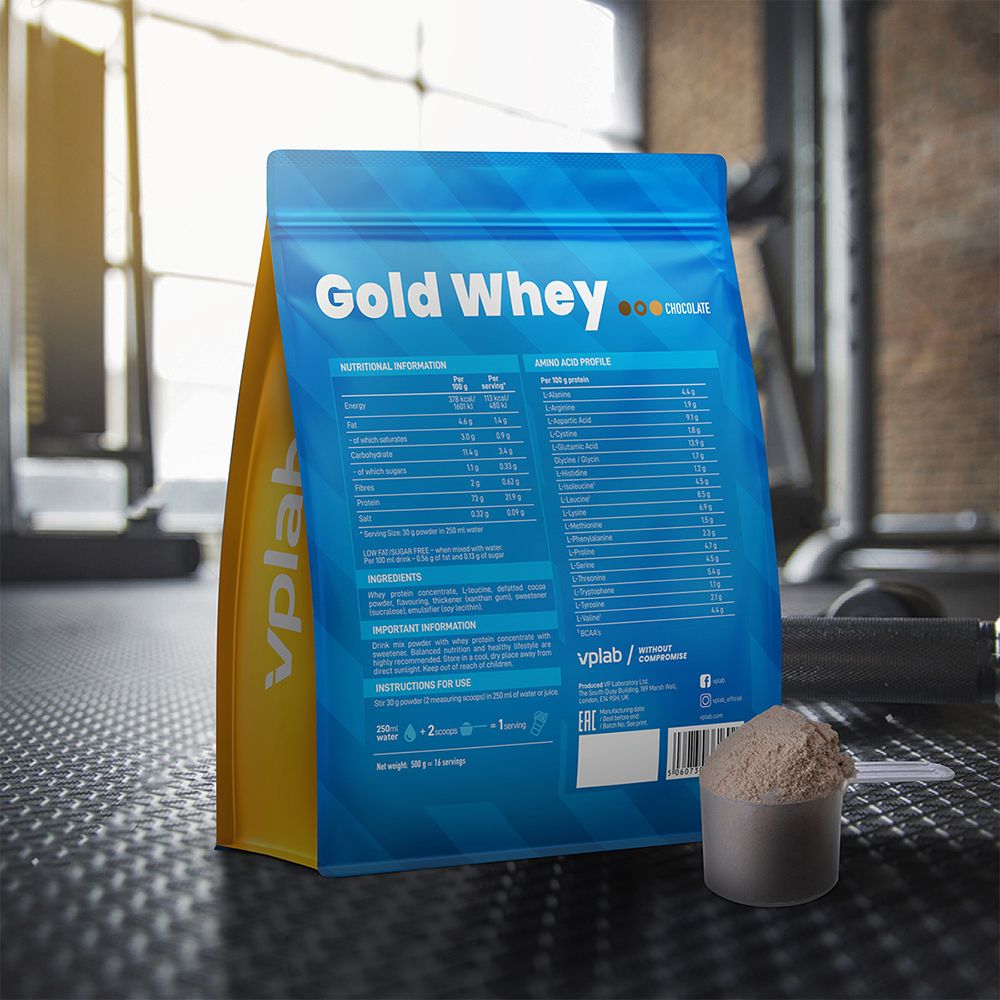 Confezione blu 'Gold Whey', misurino con polvere. Sfondo: palestra. Prodotto accanto al misurino.