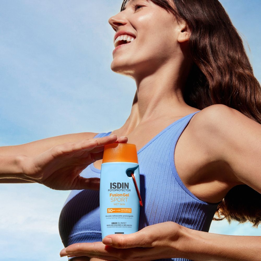Donna che tiene una bottiglia ISDIN Fotoprotettore Corpo Fusion Gel Sport SPF 50 davanti a un cielo blu.