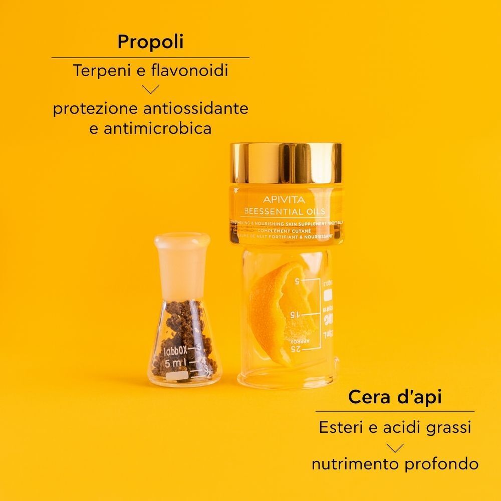 Un vasetto Apivita Beessentials Oils Balsamo Notte, un becher con propoli e una scorza d'arancia su sfondo giallo.