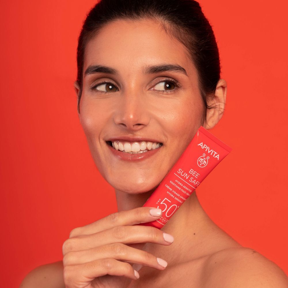 Una donna tiene un tubetto rosso di crema solare viso APIVITA SPF 50+ su sfondo rosso.