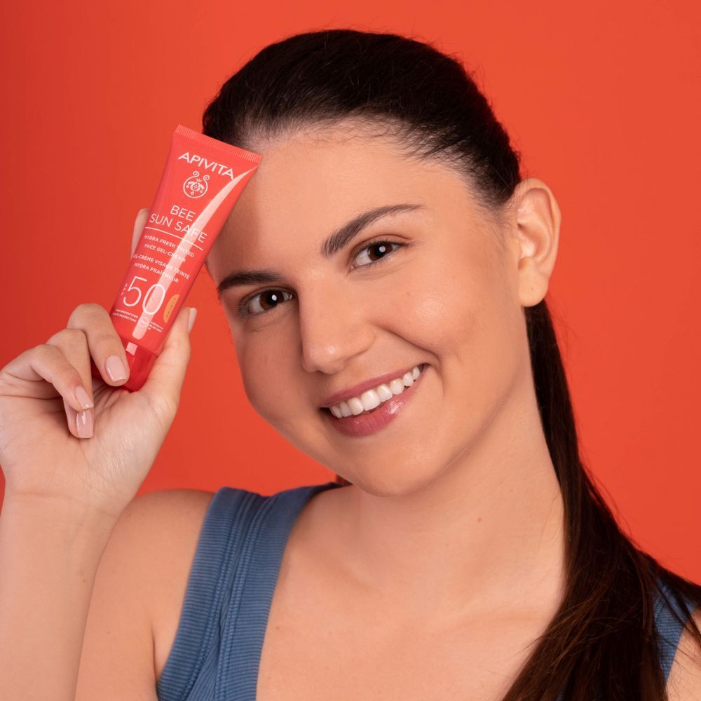 Donna sorridente tiene un tubo APIVITA Bee Sun Safe Hydra Fresh Gel-Crema Colorata SPF50 davanti al viso.
