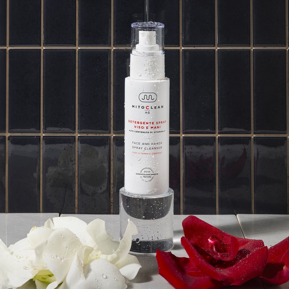 Flacone spray di detergente viso e mani Mitochon Mitoclean AG su un supporto trasparente, con fiori.