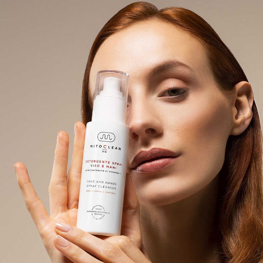 Una donna dai capelli rossi tiene in mano un flacone spray di detergente viso e mani Mitochon Mitoclean AG.
