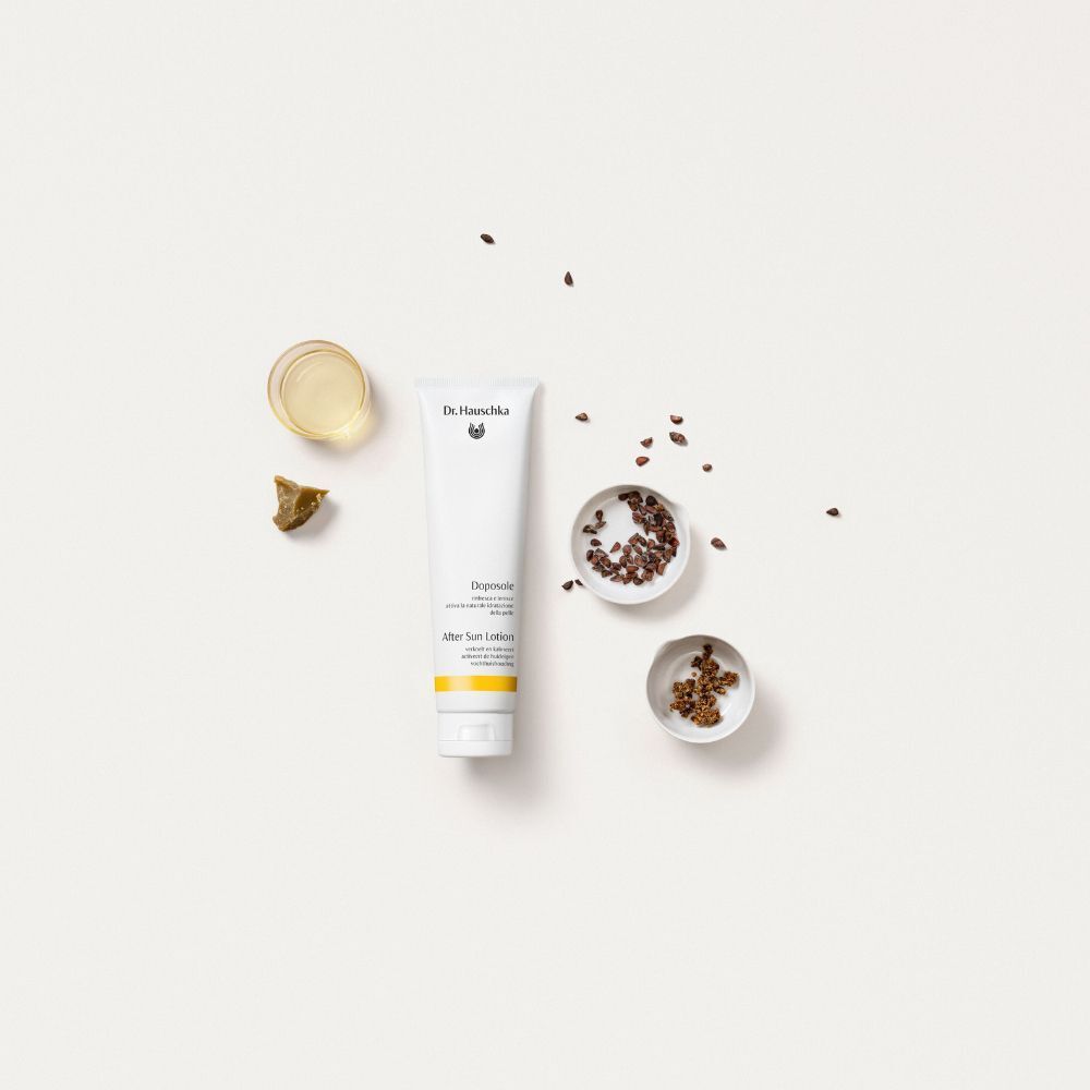 Un tubo di Dr. Hauschka Doposole After Sun Lotion accanto a piccole ciotole di ingredienti e un bicchiere d'olio.