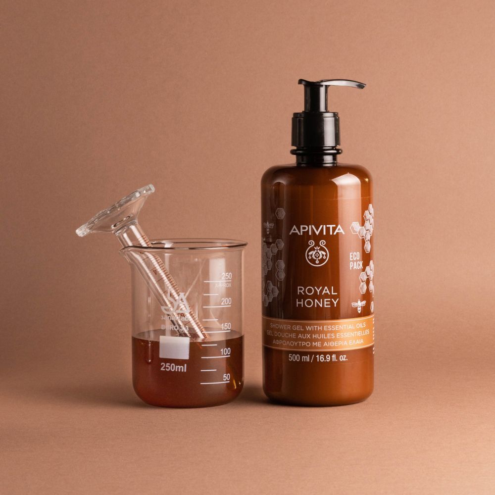 Flacone marrone di bagnoschiuma APIVITA Royal Honey accanto a un becher con miele e pipetta.