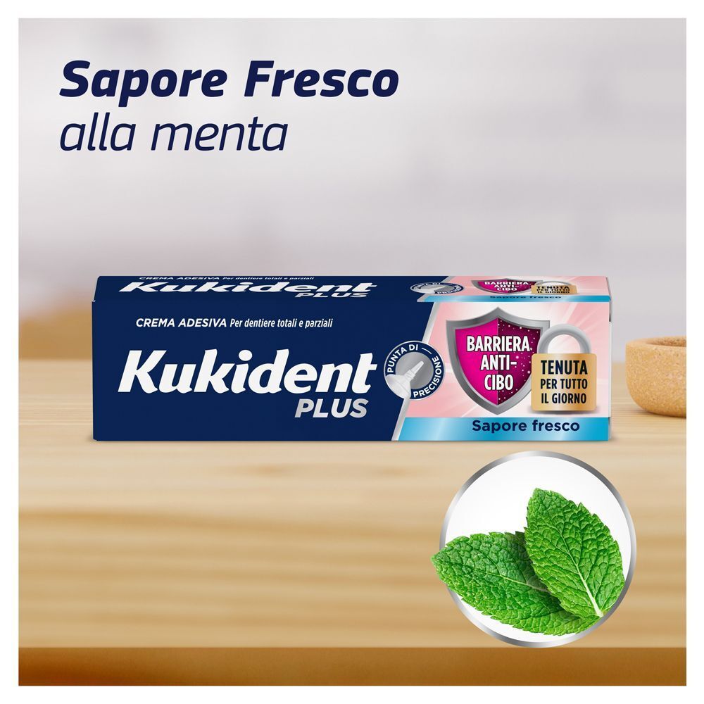 Crema adesiva Kukident Plus al sapore di menta, su tavolo in legno con foglie di menta.