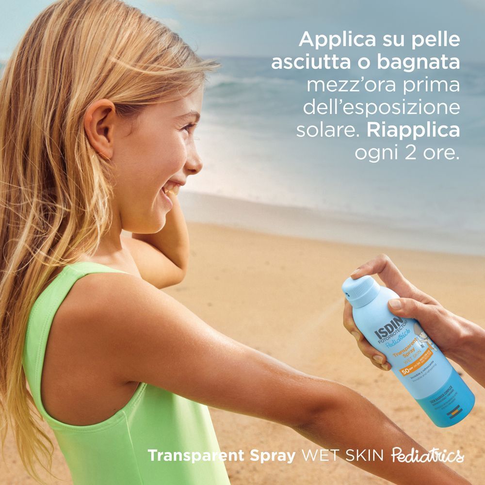 Una mano spruzza ISDIN Fotoprotector Pediatrics Transparent Spray SPF 50 sulla pelle in spiaggia.
