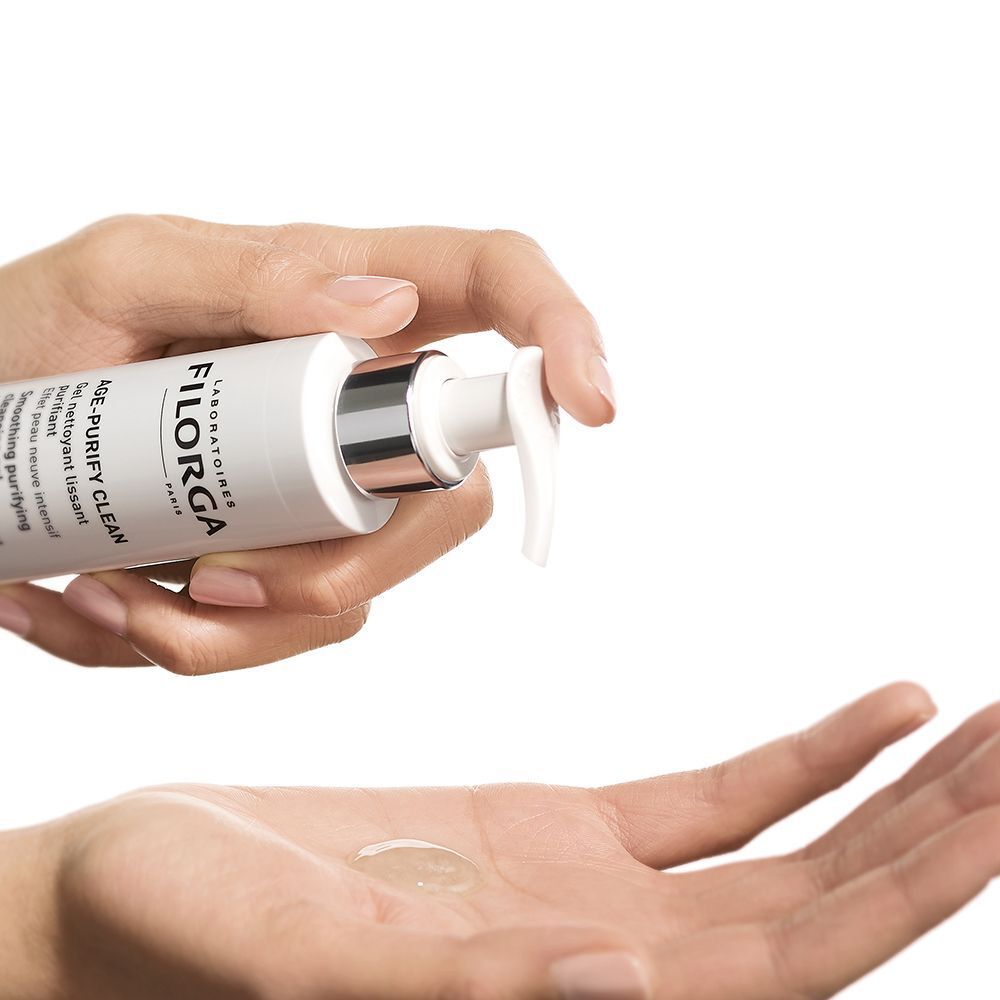 Una mano preme un flacone a pompa bianco FILORGA AGE-PURIFY CLEAN, il gel gocciola nell'altra mano.