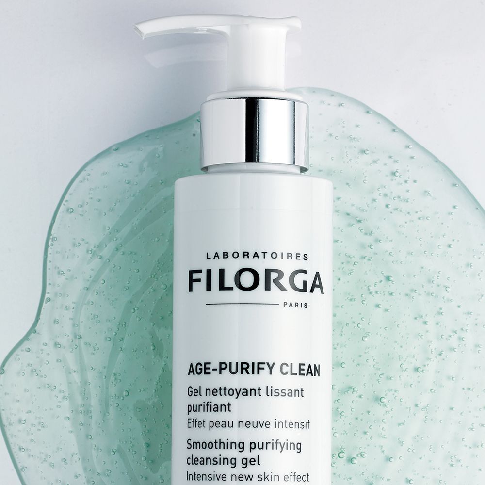 Flacone a pompa bianco FILORGA AGE-PURIFY CLEAN su una superficie verde ondulata.