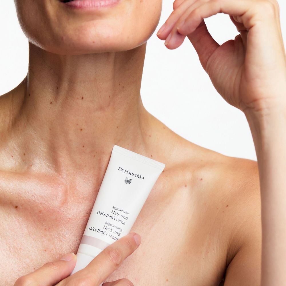 Una mano tiene un tubetto di Crema Rigenerante Collo e Décolleté Dr. Hauschka vicino al collo e décolleté di una persona.