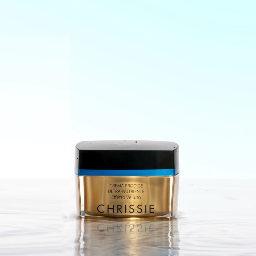 Un vasetto dorato Chrissie Crema Prodige Ultra-Nutriente Effetto Velluto su una superficie riflettente.