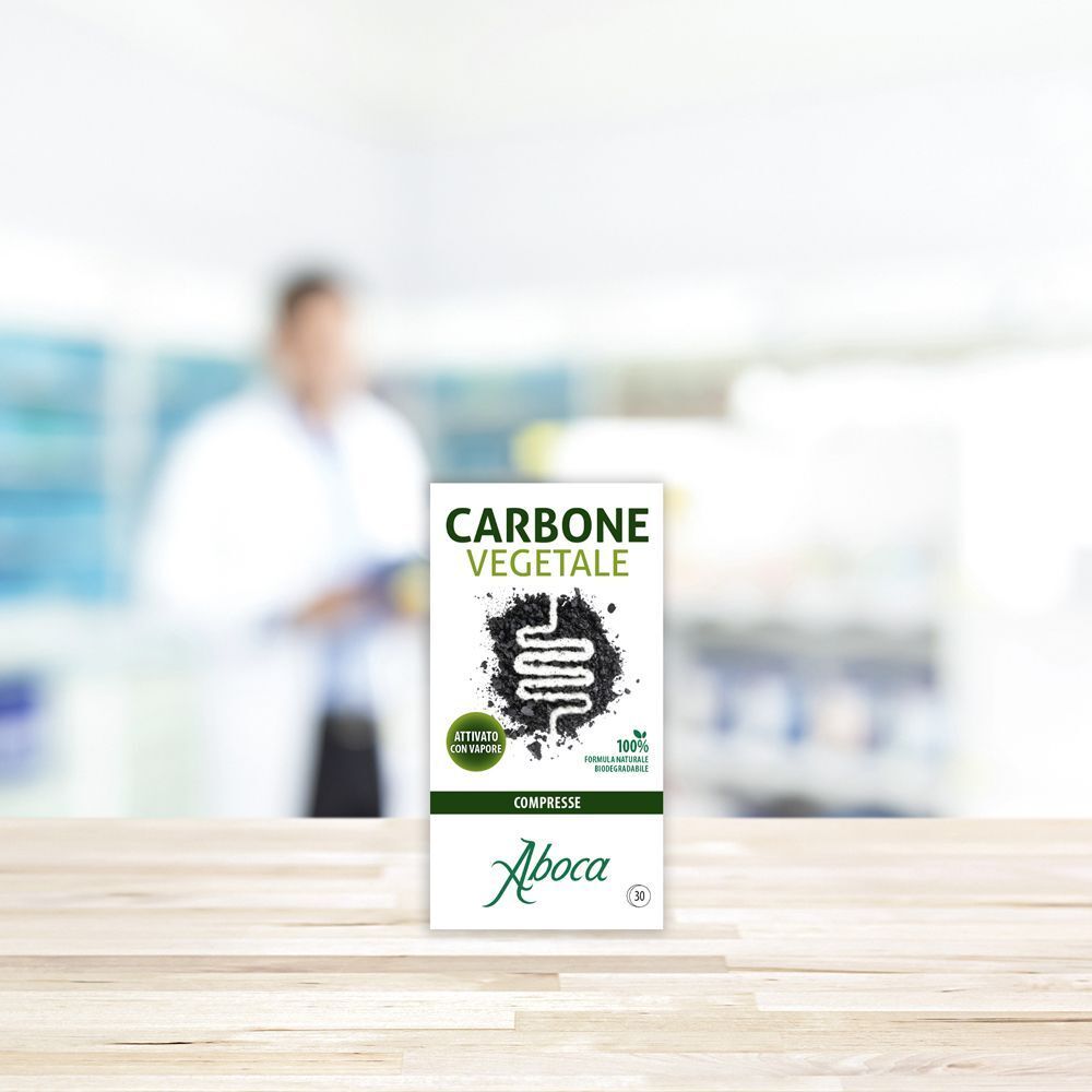 Una confezione di Aboca Carbone Vegetale compresse su un tavolo di legno, con farmacista sullo sfondo.