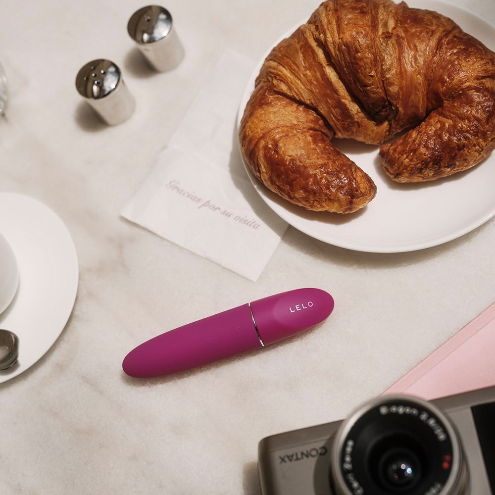 Massaggiatore personale Lelo Mia3 viola su tavolo di marmo vicino a croissant e fotocamera.