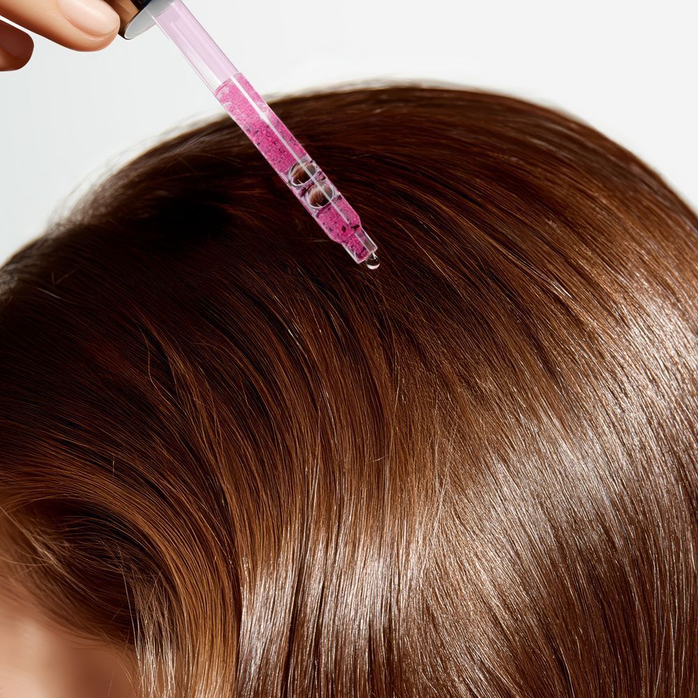 Una pipetta con siero rosa posizionata sopra capelli castani, una goccia cade.