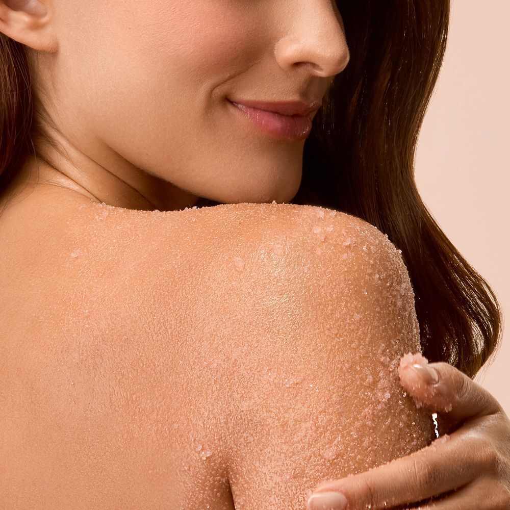 Spalla e collo di una donna con granuli di scrub rosa sulla pelle.