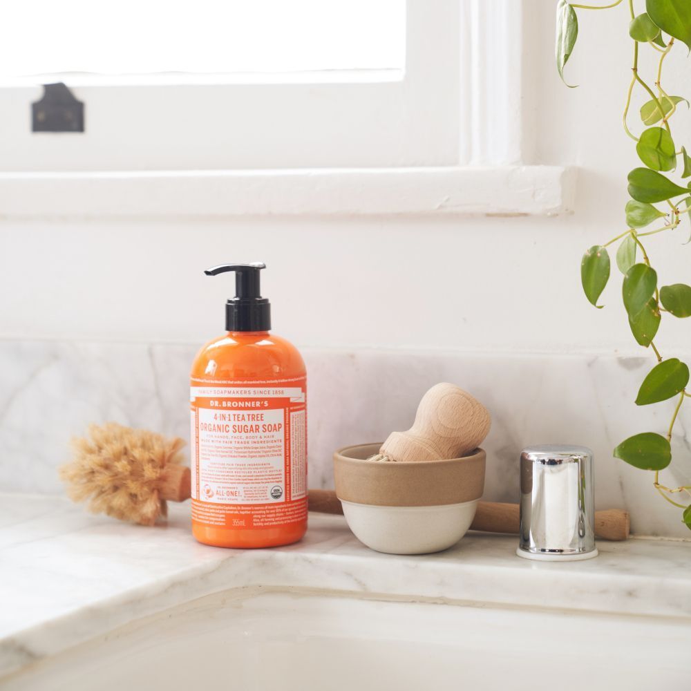Sapone biologico allo zucchero Dr. Bronner's Tea Tree accanto a una spazzola e una ciotola su un piano di lavoro in marmo.