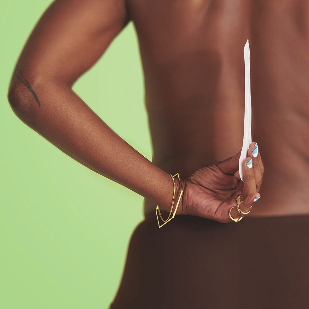 Persona tiene un prodotto bianco dietro la schiena. Sfondo verde. La persona indossa gioielli.