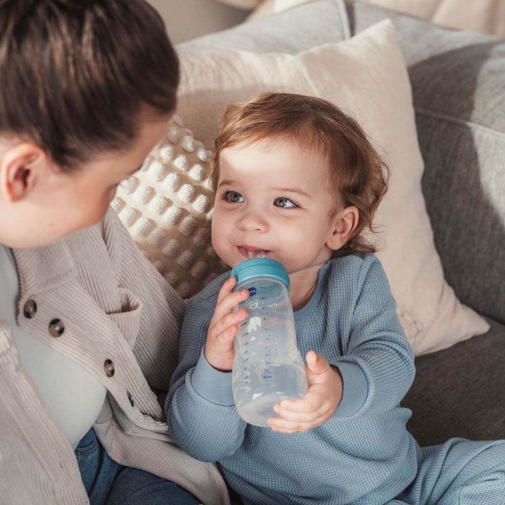 Un bambino beve da un biberon trasparente con tappo blu. Una donna tiene il biberon.