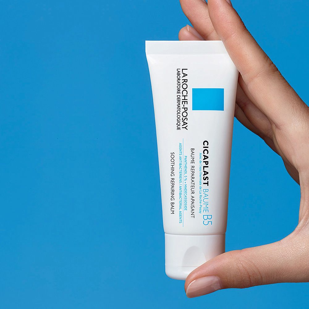 Mano che tiene il tubo La Roche-Posay CICAPLAST BAUME B5. Bianco, blu e testo. Sfondo blu.