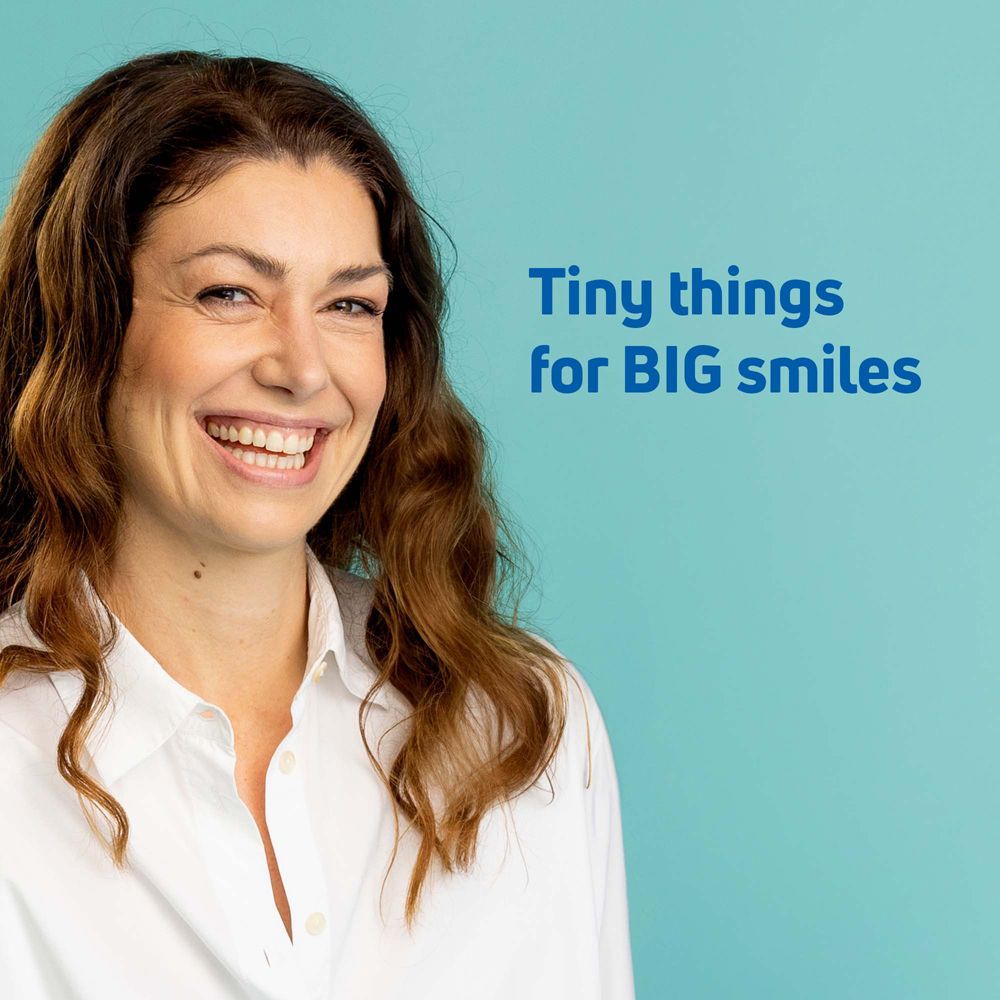 Donna sorridente con lunghi capelli castani, indossa una camicia bianca. Sfondo blu con testo "Tiny things for BIG smiles".