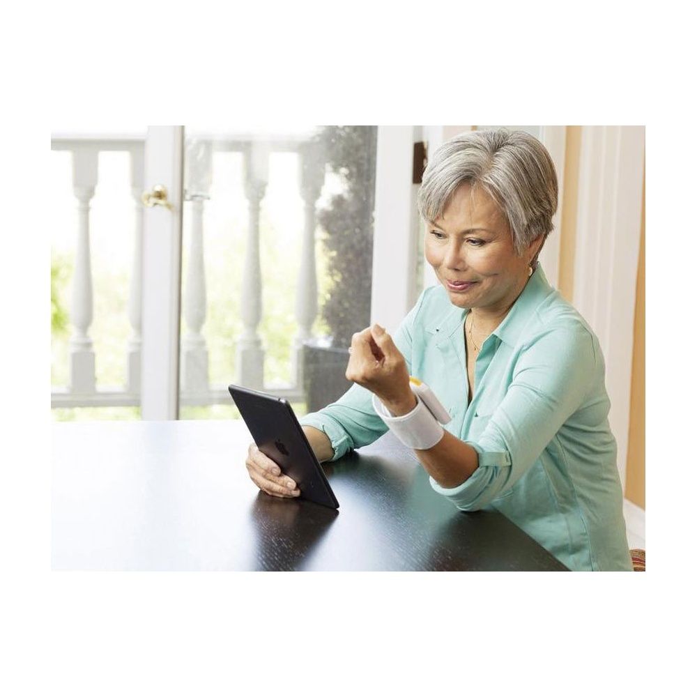 Donna anziana seduta a un tavolo che guarda un tablet. Indossa un misuratore di pressione al polso.