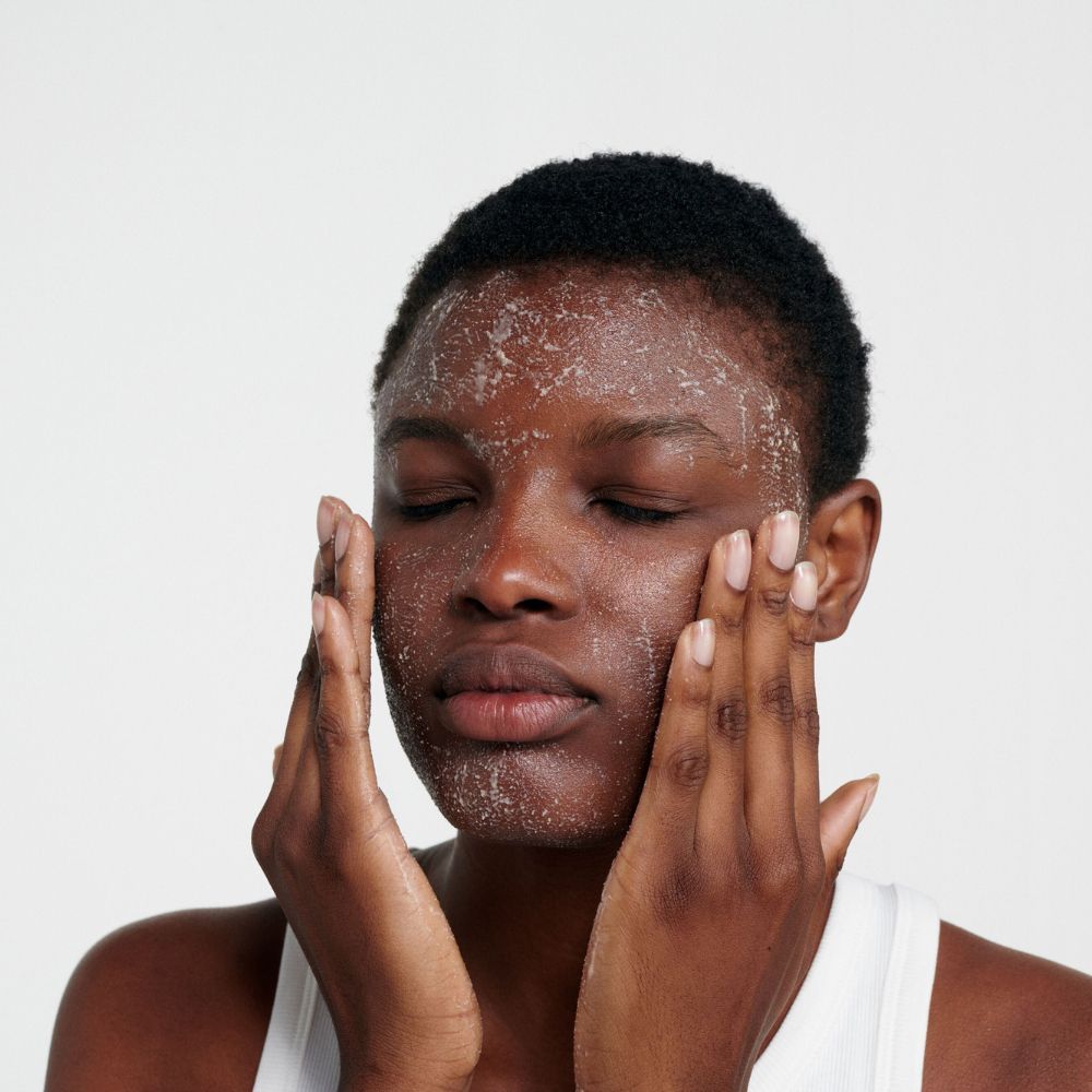Persona che si lava il viso con un prodotto color crema. Le mani strofinano il prodotto. Occhi chiusi.