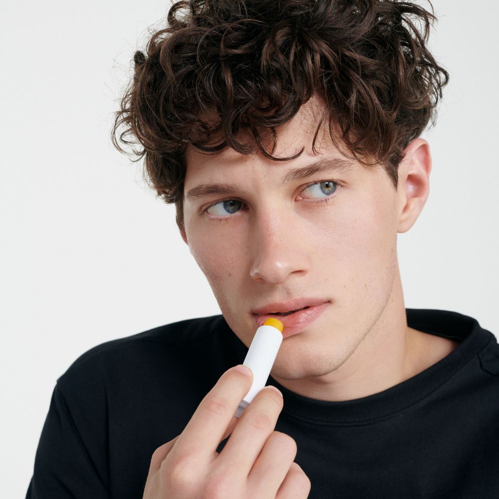 Uomo usa stick per labbra. Stick bianco con fascia gialla. Marchio: Dr. Hauschka.