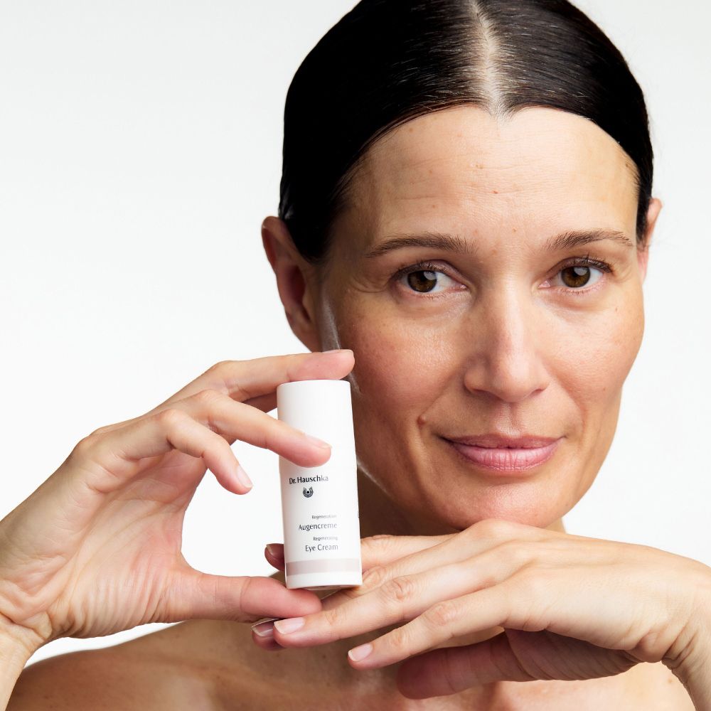 Donna tiene un tubo bianco. Scritte: Dr. Hauschka, Regeneration, Eye Cream. Donna con capelli scuri.