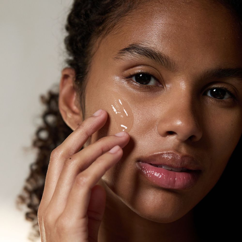 Primo piano di una donna che applica un siero trasparente sulla pelle. Viso e mano visibili.