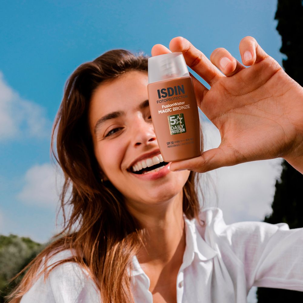 Donna che tiene in mano un flacone di crema solare. ISDIN, Fusion Water, Magic Bronze, SPF 50. Sorride. Cielo blu.