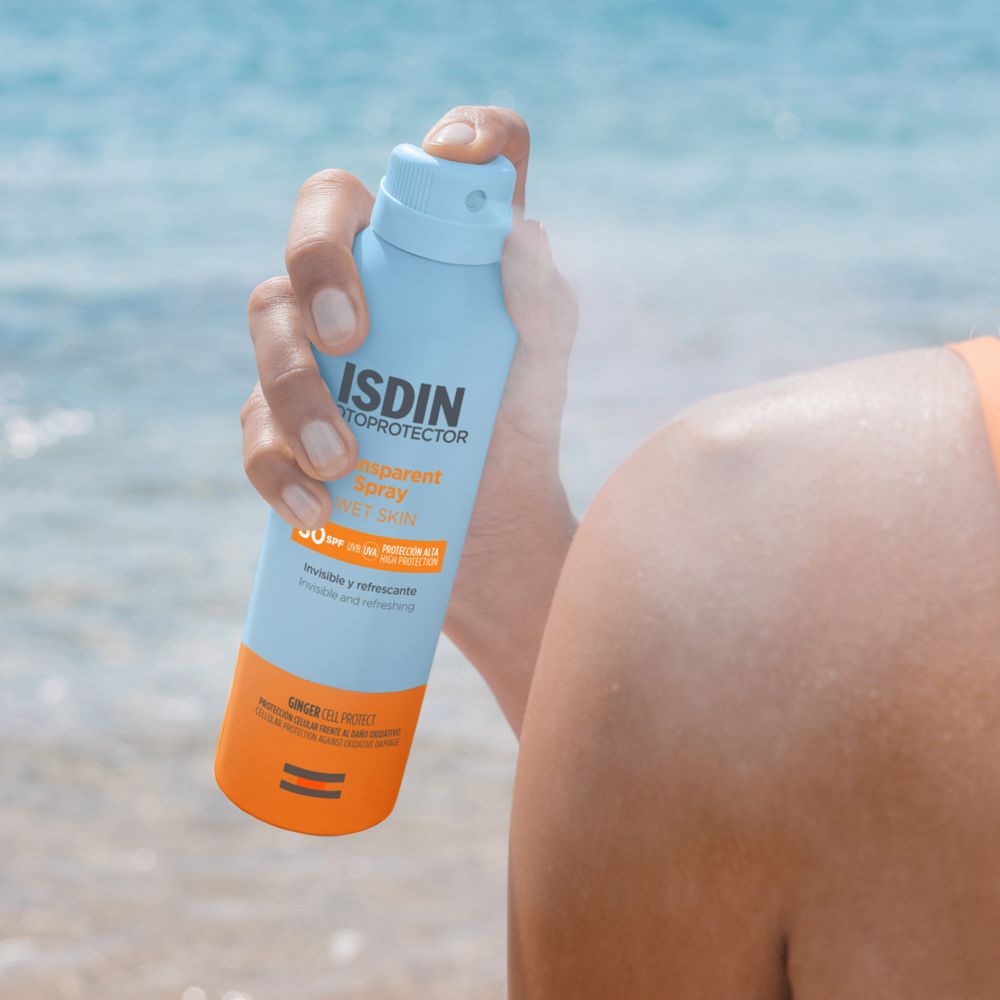 Persona che spruzza protezione solare sulla spalla. Flacone azzurro con testo: ISDIN Fotoprotector, Transparent Spray Wet Skin SPF 30.