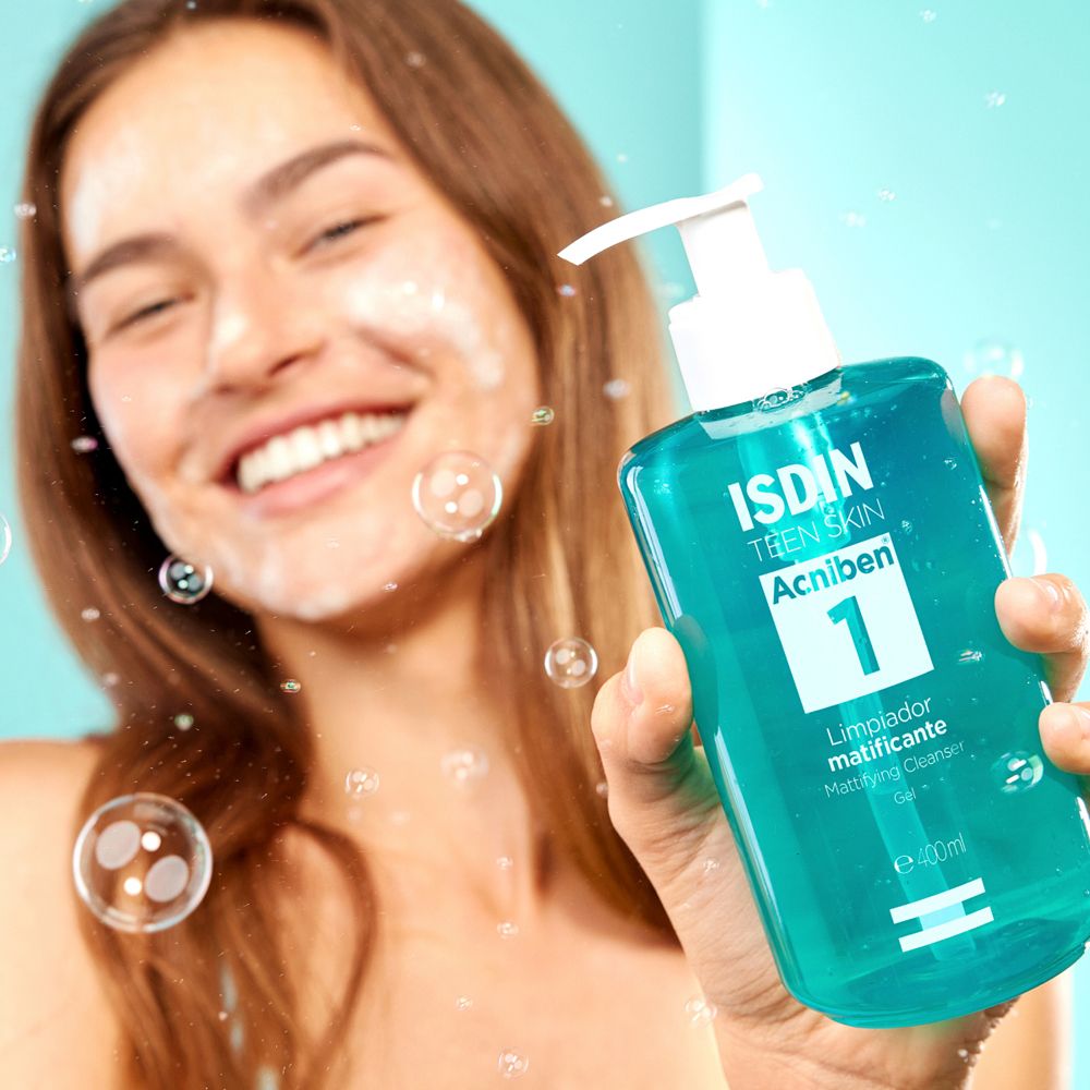 Giovane donna con schiuma sul viso tiene in mano un flacone turchese. Scritta: ISDIN Teen Skin Acniben 1.