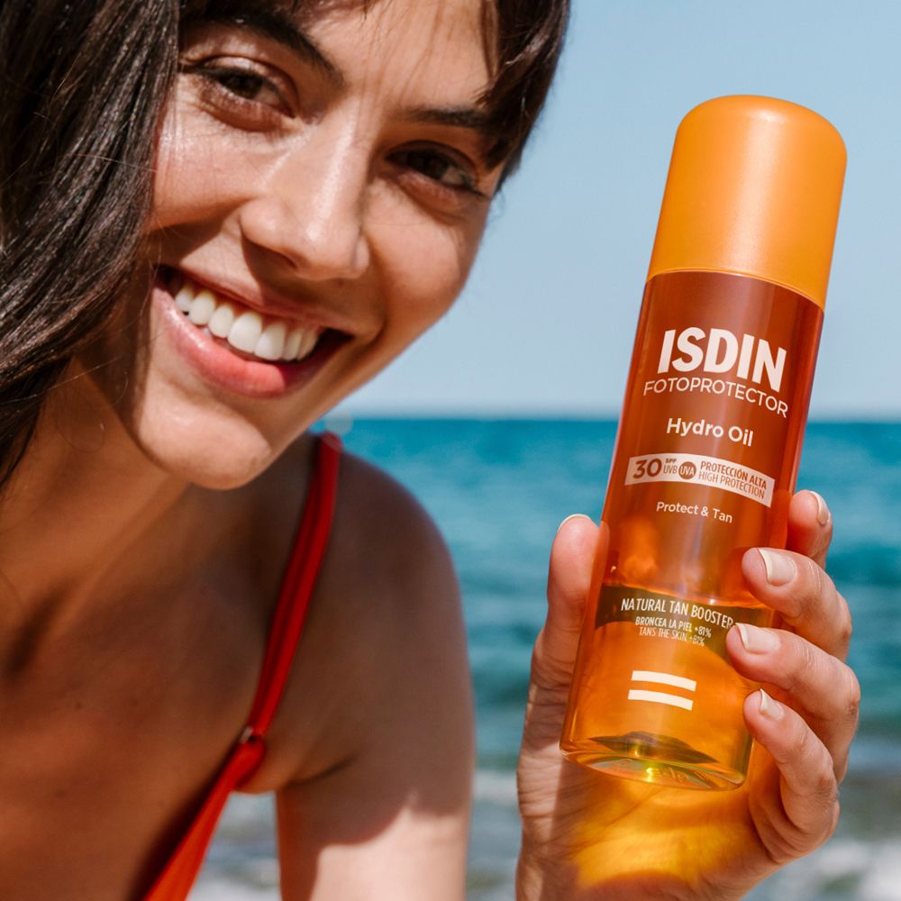 Donna tiene ISDIN Hydro Oil SPF30. Sorride in spiaggia. Flacone arancione con informazioni sul prodotto.