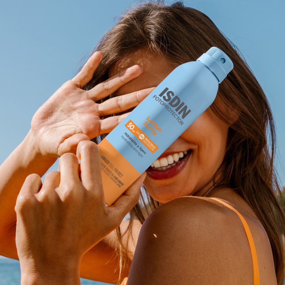 Donna tiene in mano spray blu davanti al viso. Testo: ISDIN, Lotion Spray, 50. Sfondo: mare e cielo.