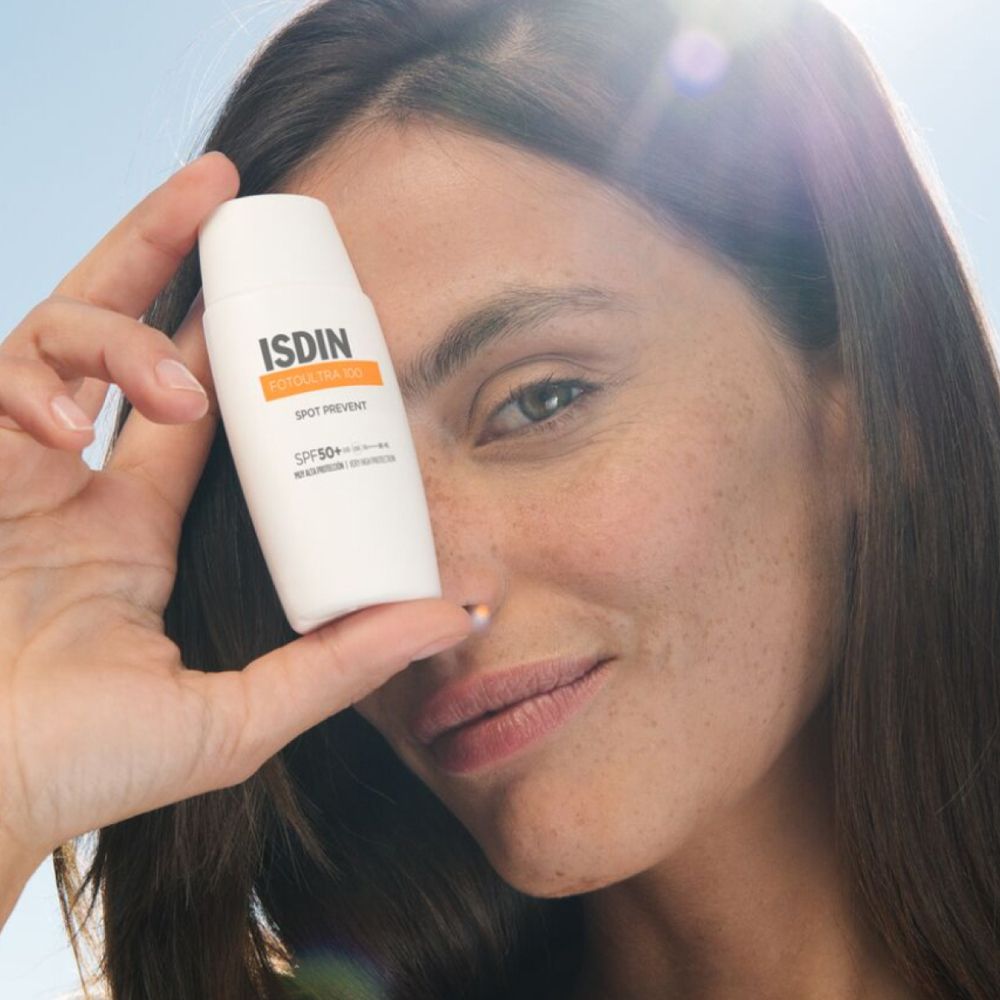 Donna tiene in mano un flacone bianco con il logo ISDIN davanti al viso. Nome del prodotto: ISDIN FOTOULTRA 100 Spot Prevent. Pelle con lentiggini.