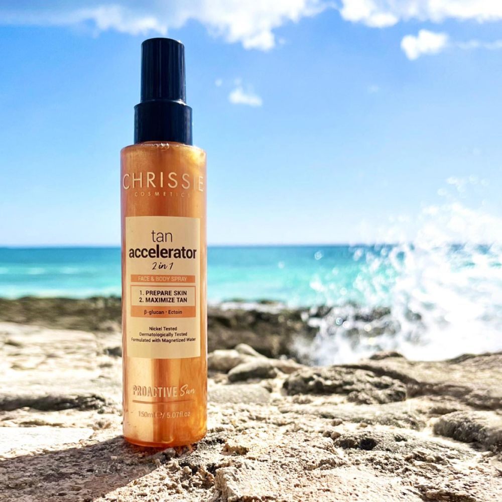 Spray acceleratore di abbronzatura Chrissie Cosmetics sulla spiaggia. Flacone marrone con erogatore nero. Mare e rocce sullo sfondo.