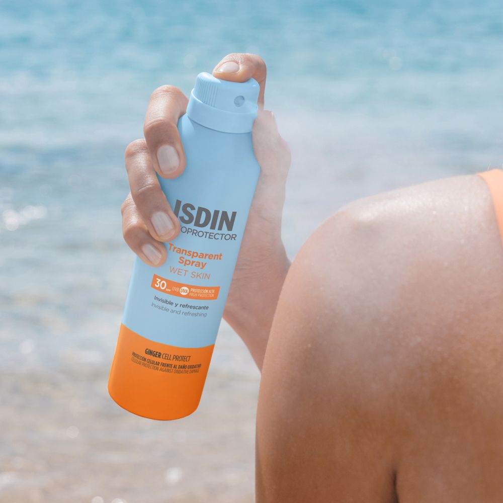 Persona che tiene un flacone spray blu. ISDIN Fotoprotector Transparent Spray Wet Skin SPF30. Spruzzato sulla pelle.