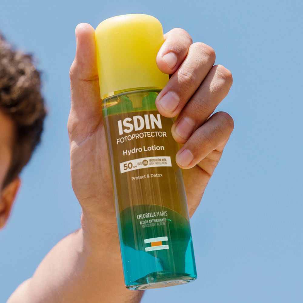 Mano che tiene ISDIN Fotoprotector Hydro Lotion. Tappo giallo, flacone trasparente. Cielo blu sullo sfondo.