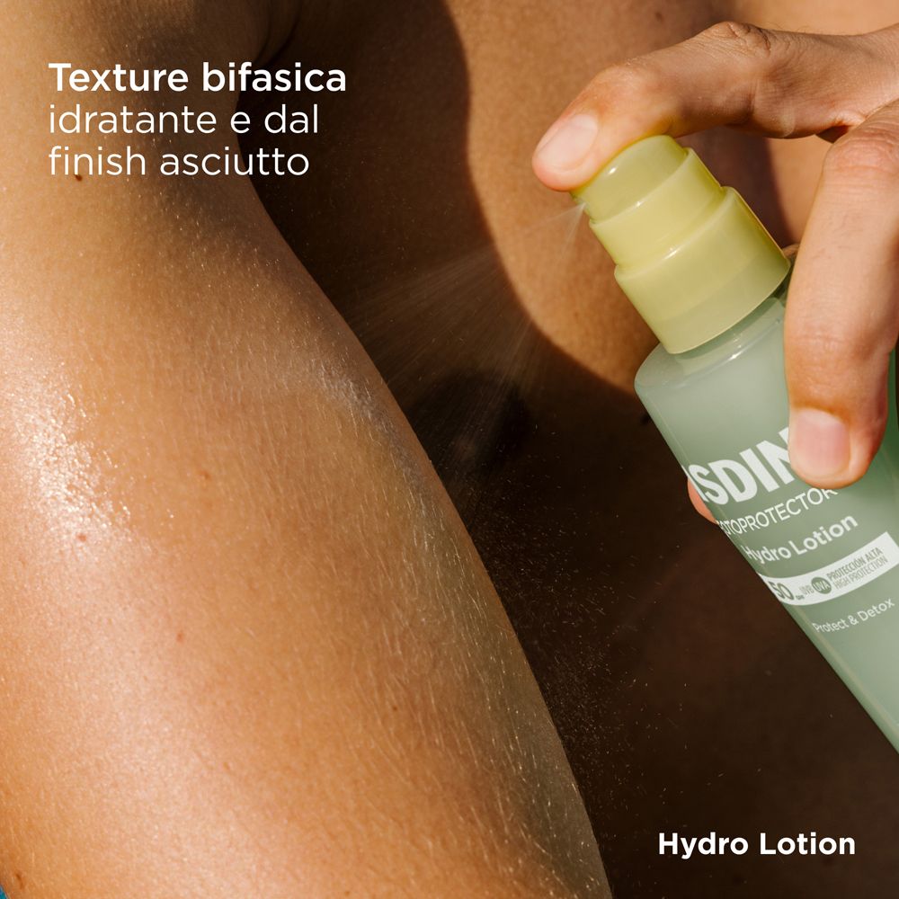Spruzzare ISDIN Hydro Lotion sul braccio. Tappo giallo, flacone verde. Testo: Texture bifasica idratante e dal finish asciutto.