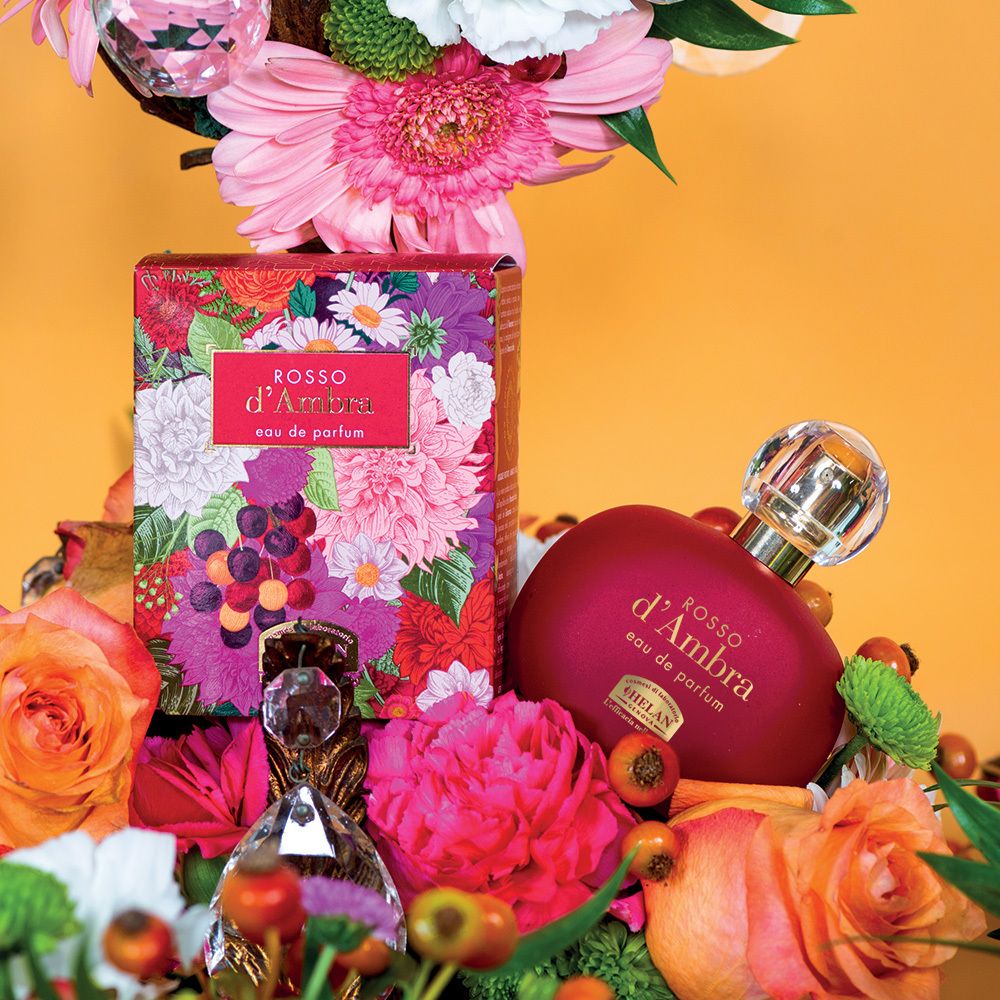 Flacone e confezione con fiori. Scritta: ROSSO d'Ambra, Eau de Parfum. Flaconi aggiuntivi. Sfondo giallo.