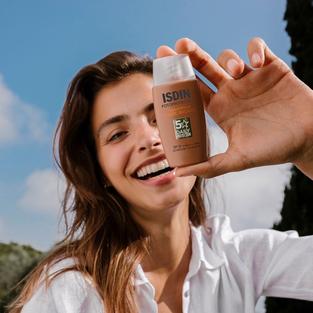 Donna tiene ISDIN Fotoprotector Fusion Water Bronze SPF50. Sorride. Sfondo: cielo e alberi.
