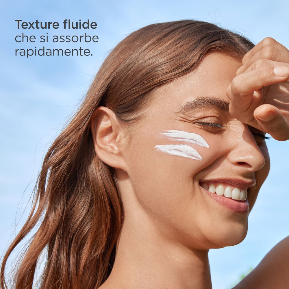 Primo piano di una donna che applica crema solare sul viso. Testo: Texture fluide che si assorbe rapidamente.