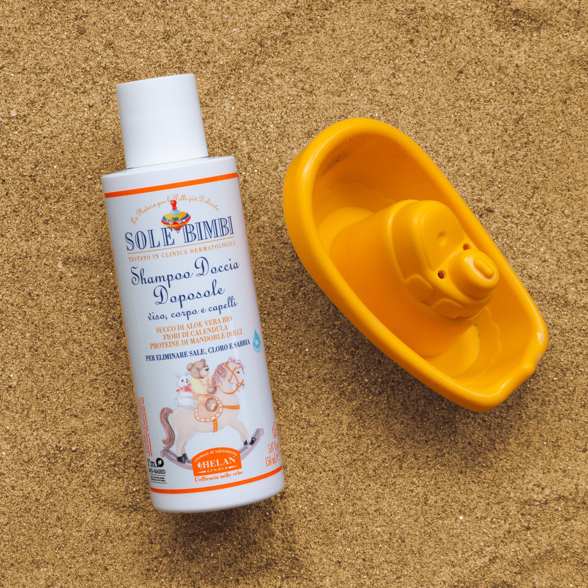 Flacone prodotto e barchetta giocattolo sulla sabbia. Testo: SOLE BIMBI Shampoo Doccia Doposole. Marca: Helan.