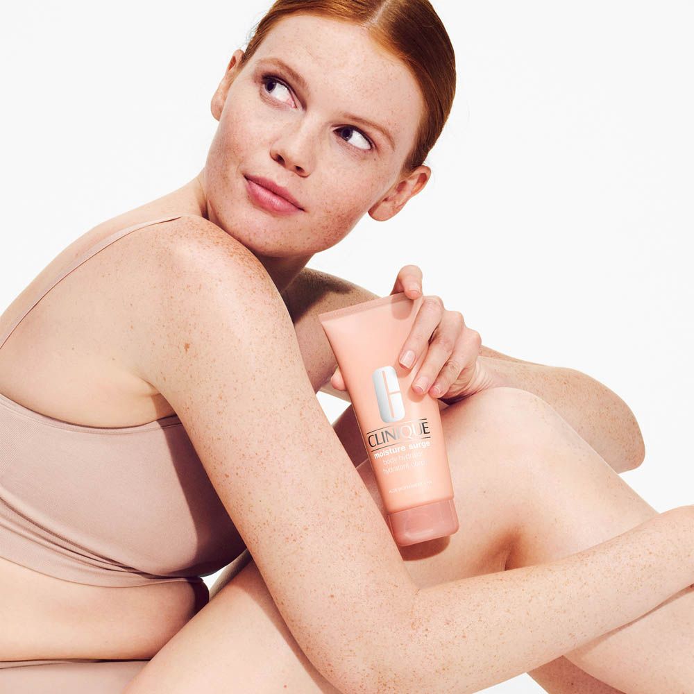 Una donna con i capelli rossi tiene in mano un tubetto rosa di Clinique Moisture Surge.