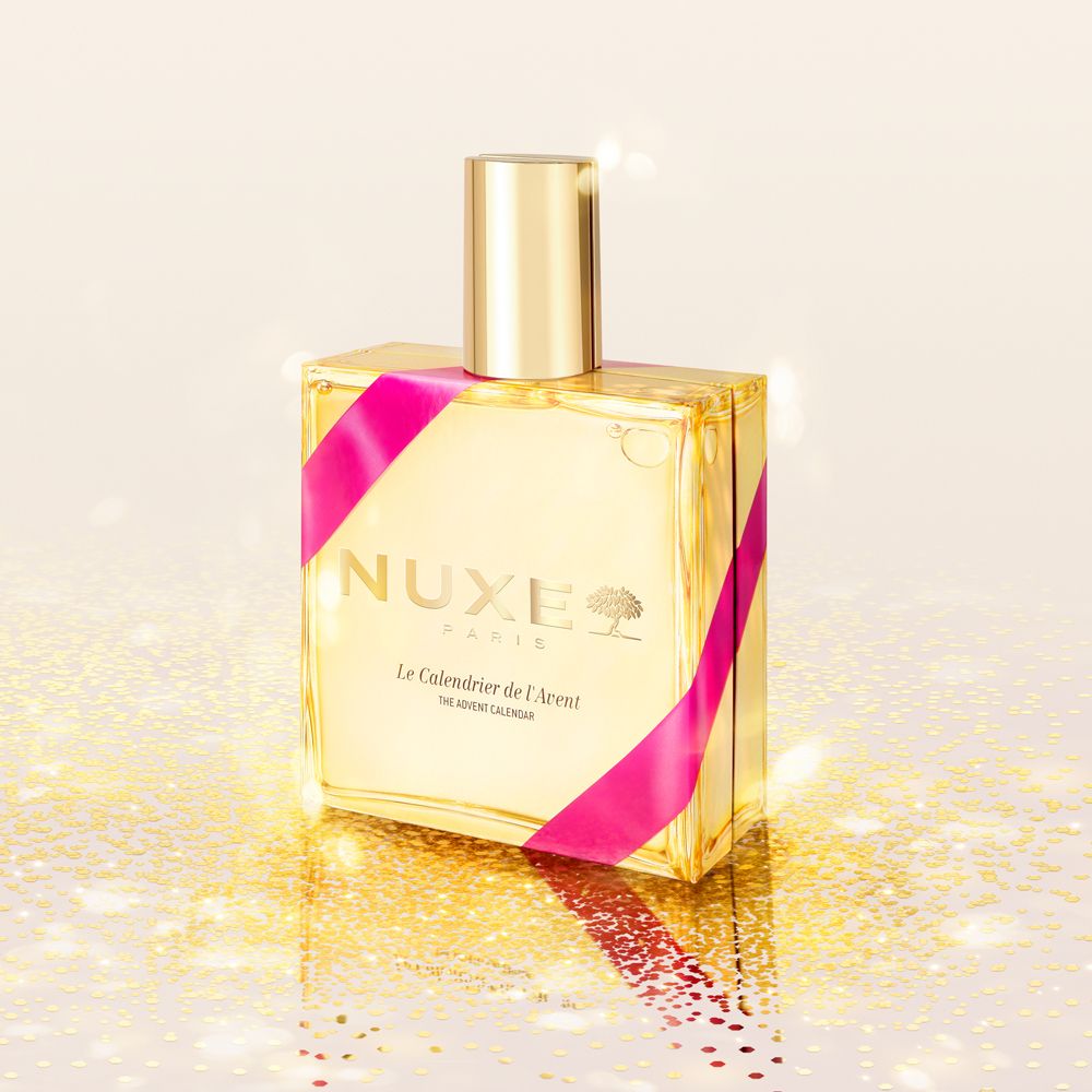 Flacone dorato con nastro rosa. Scritta: Nuxe Paris, Le Calendrier de l'Avent, The Advent Calendar.