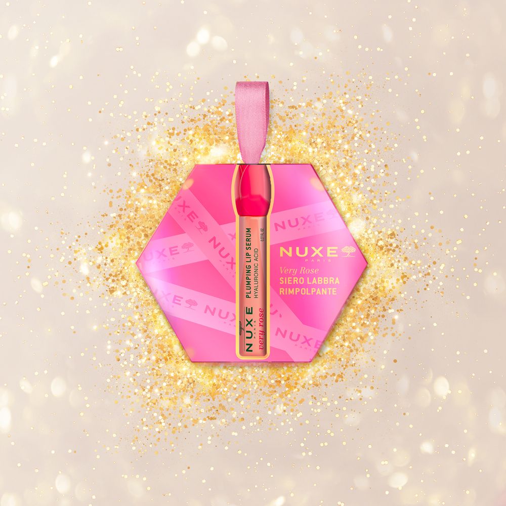 Confezione regalo esagonale rosa con fiocco rosa, circondata da glitter dorati. Contiene un siero labbra. Scritta: Nuxe, Very Rose.